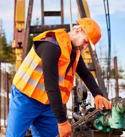 Oil man tightening lug nuts with industrial wrench in oil field. Side view of male technician using industrial wrench while repairing pipe of oil well pump jack. Oil worker in protective helmet and work vest tightening bolt on pipe. Concept of petroleum industry